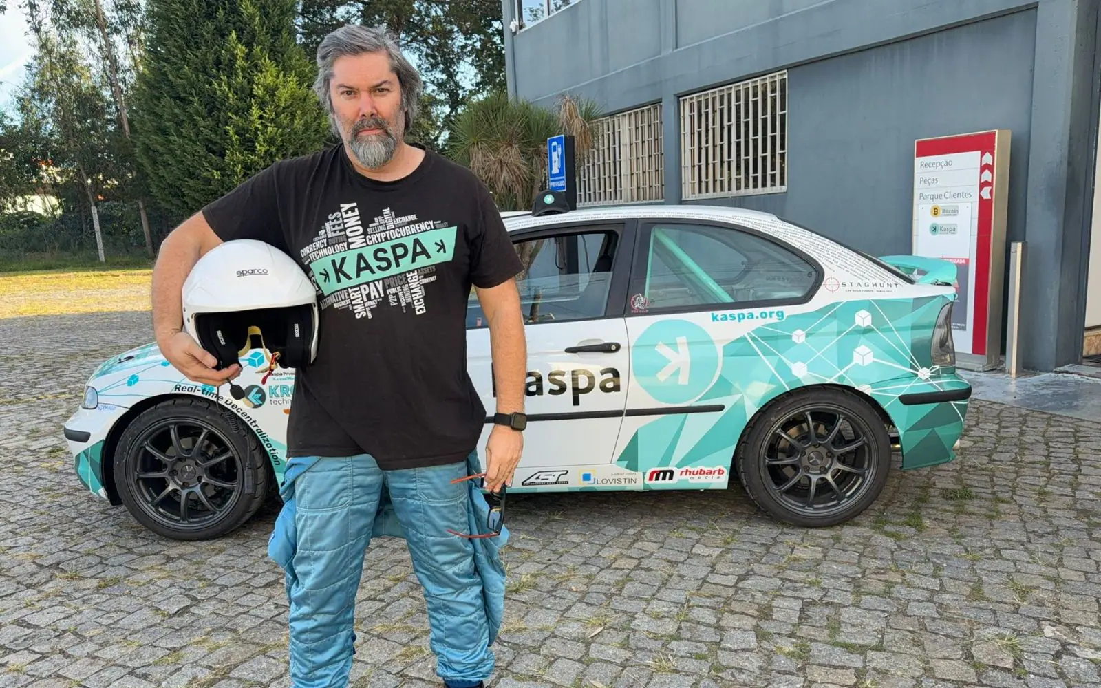 The driver in a Kaspa shirt and blue racing overalls, holding a white helmet in front of the finished rally car