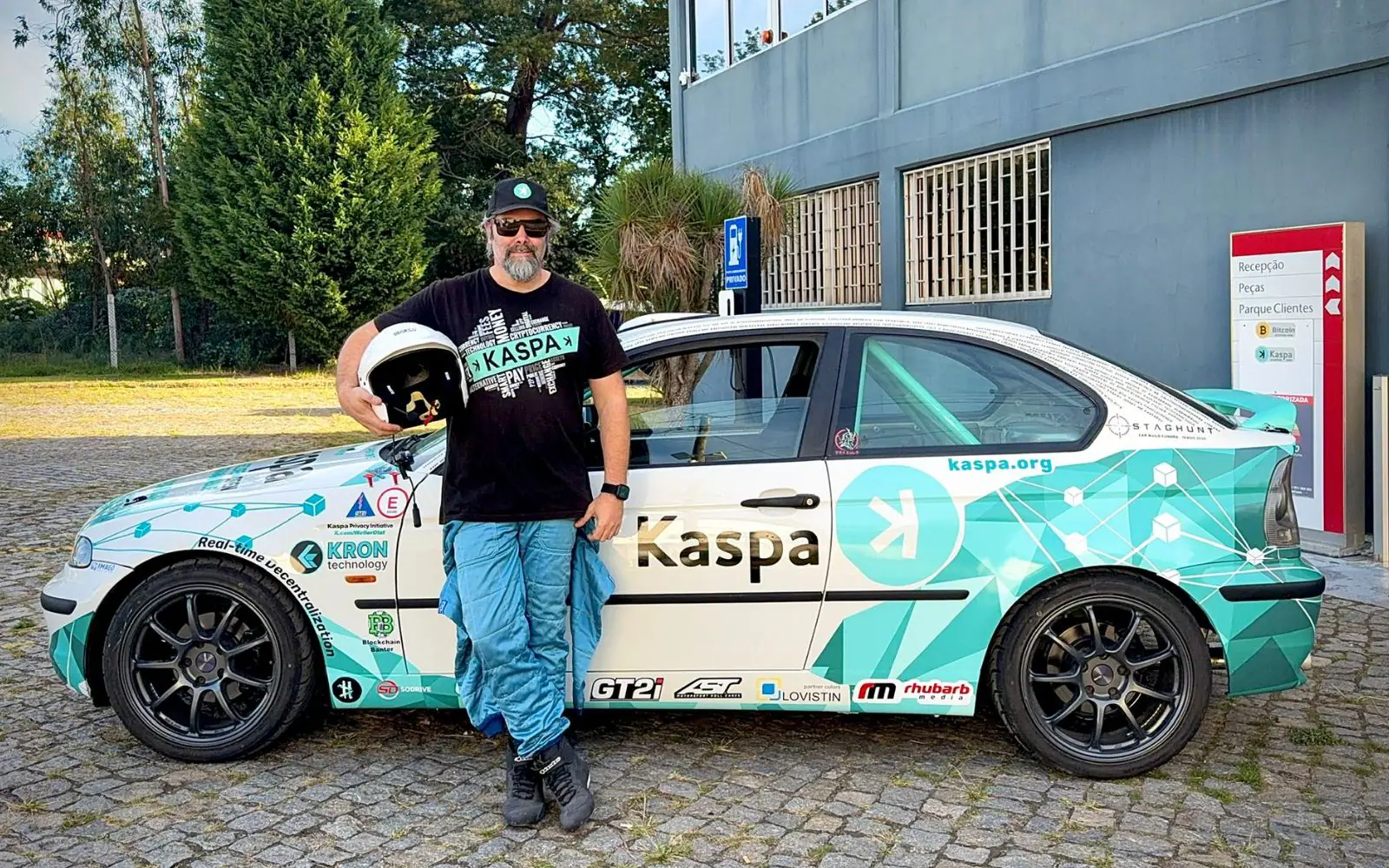 The driver in a cap and sunglasses leaning on the finished rally car, helmet in hand