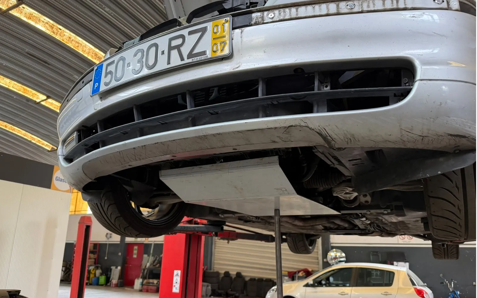 The silver BMW raised on a two-post lift in the workshop, seen from below the front bumper with the undertray and suspension exposed