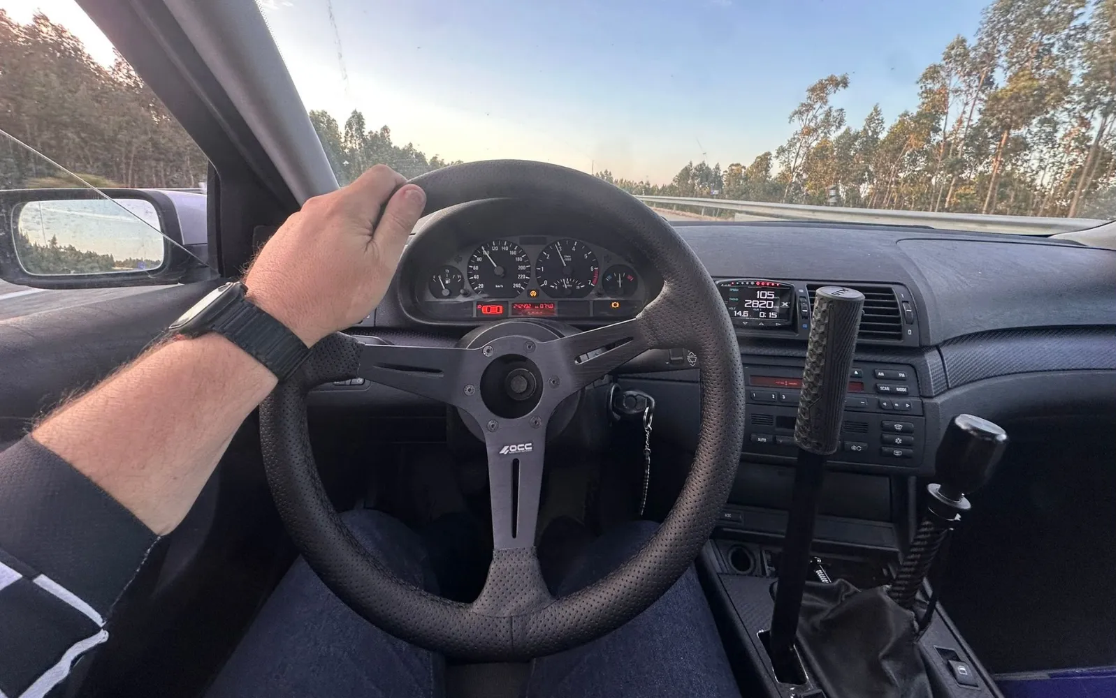 Driver's view over an OCC steering wheel and the BMW dials on a eucalyptus-lined road at dusk