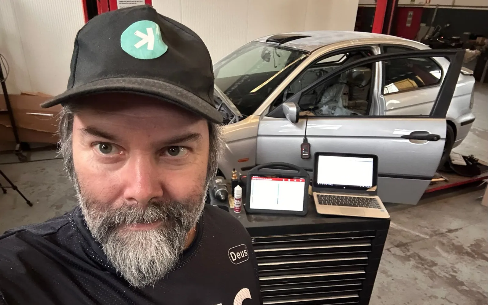 The driver beside the silver BMW on the lift, with a diagnostic tablet and a laptop set up on a tool chest