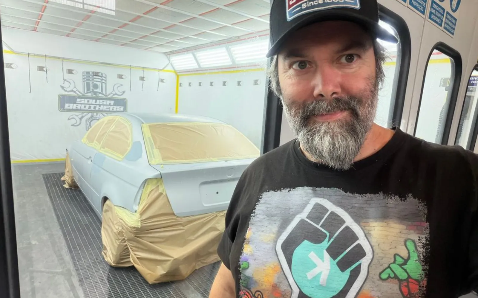 The driver inside the spray booth beside the primed and masked shell, before colour goes on