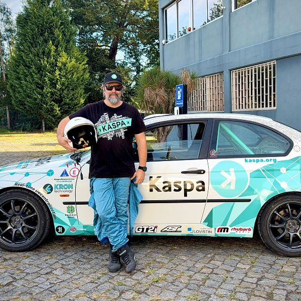 Extriage standing beside the white and teal Kaspa Rally car, holding a white helmet