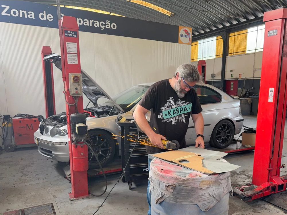 A mechanic in a Kaspa T-shirt grinding a metal body panel, sparks flying, with the rally car's silver shell raised on a two-post lift behind them in the Auto Souto workshop